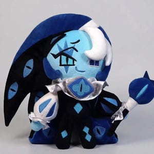 May include: A plush doll of a character with a light blue face, white hair, and a dark blue hat and outfit. The doll has a staff with a blue orb and star. The character's outfit includes a silver collar and diamond accents.
