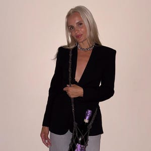 May include: A black blazer, grey trousers, and a silver chain necklace. A dark grey macrame bottle holder with a long strap carries a bottle of sparkling wine with a purple foil top. The bottle holder features a knotted net design with fringe.