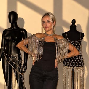 May include: A silver-toned metallic fringe shoulder piece featuring a high-neck collar design with cascading strands of reflective beads. The accessory is worn over a black top and is displayed alongside two mannequins wearing similar fringe garments.