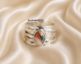 Puede incluir: Un anillo ancho de plata con una banda martillada y texturizada, y una piedra central ovalada con patrones jaspeados en tonos blanco, naranja y azul. El texto Jewelryhub2026 es visible sobre el anillo. El metal tiene un acabado pulido.
