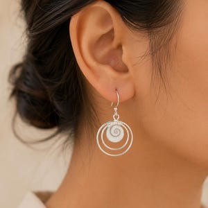May include: A silver dangle earring featuring two concentric circular hoops and a small, white, spiral-shaped shell charm suspended from a hook wire. The earring is crafted from polished silver-toned metal and displays a clean, circular design.