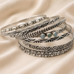 Puede incluir: Un conjunto de seis brazaletes de metal plateado con varios patrones en relieve, incluyendo motivos florales, formas geométricas y una fila de pájaros. Un brazalete está decorado con tres pequeñas piedras cabujón redondas e iridiscentes.