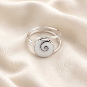 May include: A silver ring featuring a circular white shiva eye shell inlay with a natural dark grey spiral pattern. The band is constructed from a thin, polished silver wire that wraps around the finger, creating an open, layered design.