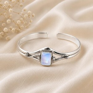 May include: A silver cuff bracelet featuring a central rectangular moonstone cabochon. The metal band displays a polished finish with decorative swirling wire details and small spherical accents on either side of the stone setting.