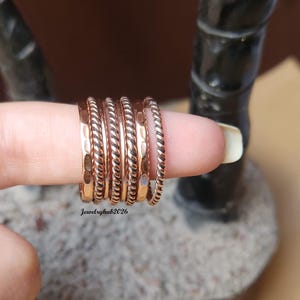May include: A selection of copper-coloured rings in diverse designs, including hammered and twisted textures. The rings are stacked on a finger, displaying their different styles. The jewellery is presented against a blurred background.