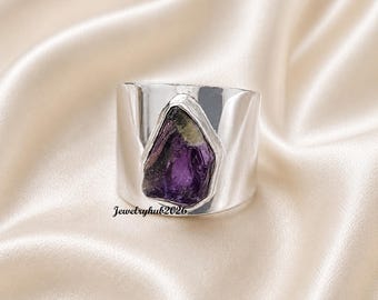 Puede incluir: Un anillo de banda ancha de plata con una piedra preciosa de amatista púrpura cruda e irregular engastada en el centro. La superficie del metal es pulida y reflectante. El texto Jewelryhub2026 está impreso en negro en la base.