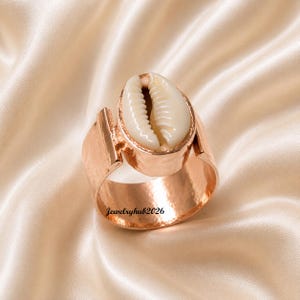 May include: A rose gold-toned ring featuring a central white cowrie shell set within a smooth metal bezel. The band has a wide, flat, and hammered-texture design. The text Jewelryhub2026 is printed in black on the lower portion of the ring band.