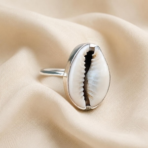 Peut inclure: Une bague en argent avec un coquillage cauri blanc comme pièce centrale. Le coquillage présente une surface lisse et brillante avec une ouverture dentelée sombre au centre. L'anneau en métal est en argent poli, maintenant le coquillage.