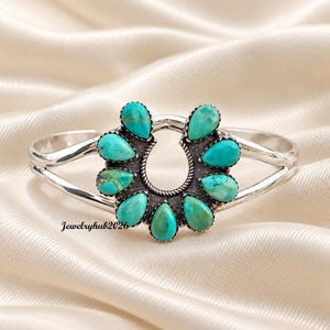 May include: A silver cuff bracelet featuring a horseshoe-shaped centrepiece adorned with nine teardrop-shaped turquoise stones. The metal frame has a polished finish and a rope-textured border around the stones. The text Jewelryhub2026 is visible.