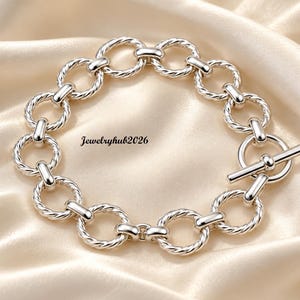 May include: A silver-toned chain bracelet featuring a series of interconnected circular links with a twisted rope texture. The bracelet includes a toggle clasp closure and the text Jewelryhub2026 is visible near the center of the circular arrangement.