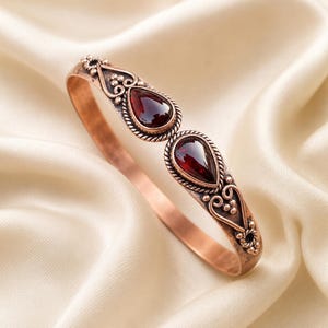 May include: A copper cuff bracelet featuring two teardrop-shaped dark red gemstones set in twisted metal borders. The metal band displays intricate scrollwork and small bead accents surrounding the stones, creating a detailed metallic finish.
