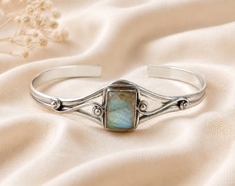 Puede incluir: Una pulsera tipo brazalete de plata con una piedra preciosa de labradorita rectangular en el centro. La banda de metal presenta un acabado pulido con detalles decorativos de alambre en espiral y pequeños acentos esféricos a ambos lados.