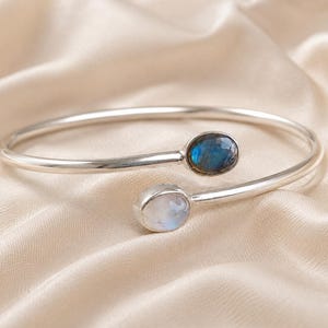 Op de afbeelding: Een zilveren open armband met twee ovaalvormige edelstenen aan de uiteinden. De ene steen is een donkere, iriserende labradoriet met blauwe flitsen, en de andere is een doorschijnende, melkachtige witte maansteen met een zachte parelmoerglans.