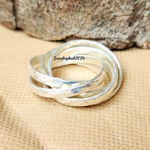 May include: A silver-coloured Russian-style ring consisting of three interlocking bands with a hammered, textured finish. The metal surface reflects light, creating highlights across the curved edges of the bands. The text Jewelryhub2026 is visible.