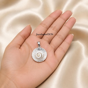 Puede incluir: Un colgante circular de plata con una incrustación central de concha blanca en espiral, rodeado por un patrón de rayos de líneas de plata grabadas. El colgante incluye una pequeña anilla de plata. El texto Jewelryhub2026 es visible.