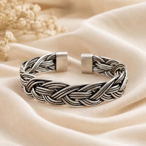 Könnte beinhalten: Ein silberfarbenes Armband in Manschettenform mit einem komplexen geflochtenen Design und polierter Oberfläche. Das Armband hat eine offene Struktur mit glatten, rechteckigen Metallkappen an beiden Enden, die ein detailliertes gewebtes Metallmuster zeigen.