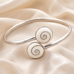 May include: A silver-toned bypass bracelet featuring two circular white shell accents. Each shell is inlaid with a brown spiral pattern, creating a distinct contrast against the polished metallic band that curves to meet the two circular focal points.