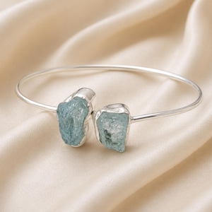 May include: A silver cuff bracelet featuring two raw, light blue aquamarine stones set at the open ends. The metal band is smooth and polished, contrasting with the textured, irregular surfaces of the two natural gemstone crystals.