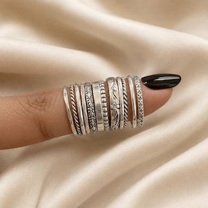 May include: A collection of eleven silver-toned metal rings arranged in a row. The rings feature various textures and patterns, including twisted rope designs, smooth bands, beaded edges, and intricate floral engravings, all displayed together.