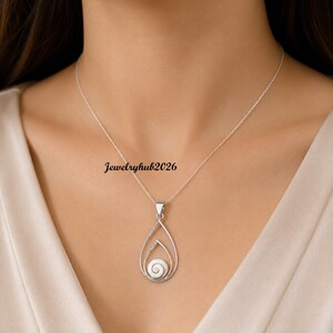 Puede incluir: Un collar de tono plateado con un colgante en forma de lágrima que encierra una pequeña concha blanca en espiral. El colgante cuelga de una delicada cadena plateada. El texto Jewelryhub2026 es visible cerca del colgante en la cadena.