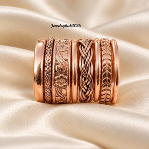 Peut inclure: Un ensemble de sept bagues couleur or rose présentant diverses textures et motifs, incluant un motif floral, un design tressé et des gravures géométriques. Le texte Jewelryhub2026 est visible au-dessus des bagues alignées en rangée.