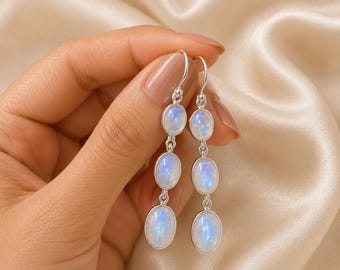 May include: A pair of drop earrings featuring three vertically aligned oval moonstones set in polished silver. Each earring hangs from a simple silver hook, showcasing the iridescent blue glow of the smooth, cabochon-cut stones in a linear design.