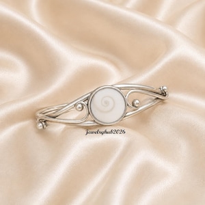 May include: A silver cuff bracelet featuring a central circular white shiva eye shell with a natural spiral pattern. The metal band has curved wire detailing and small spherical accents on each side. The text Jewelryhub2026 is visible below.
