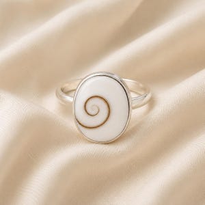 Puede incluir: Un anillo de plata con una incrustación ovalada de concha de ojo de Shiva. La incrustación es lisa, blanca y muestra un patrón de espiral marrón natural centrado en la superficie. El aro es de plata pulida y se une al engaste ovalado.