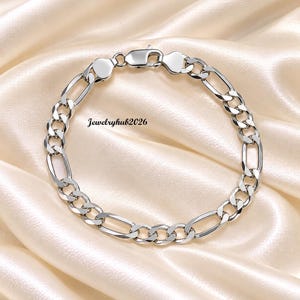 May include: A silver-toned Figaro chain bracelet featuring a series of three small circular links followed by one elongated oval link. The bracelet includes a secure lobster claw clasp and displays the text Jewelryhub2026 in the centre.