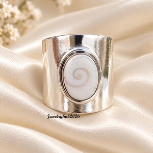 May include: A wide silver-toned band ring featuring a central oval-shaped white shiva shell inlay with a natural spiral pattern. The metal surface is smooth and reflective. The text Jewelryhub2026 is printed in small black letters near the base.