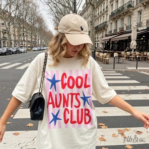 May include: A beige baseball cap with a black logo and a white t-shirt with the text "COOL AUNTS CLUB" in red and blue. The shirt has pink and white stripes. A black purse with a chain strap is also visible.