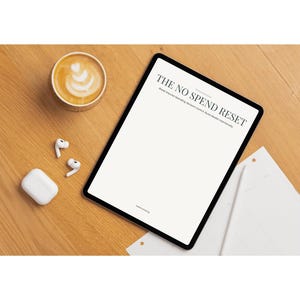 May include: A digital tablet with the text "THE NO SPEND RESET" on the screen, next to a cup of coffee with latte art, a pair of wireless earbuds, and a charging case. The items are on a wooden surface.