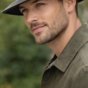Könnte beinhalten: Ein schwarzer Fedora-Hut mit dunklem Band und breiter Krempe. Der Hut wird von einer Person getragen, die eine olivgrüne Jacke trägt. Der Hintergrund ist verschwommen, mit grünem Laub.