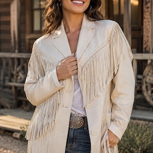 May include: A beige blazer featuring long fringe detailing across the chest, shoulders, and lower hem. The jacket has a notched lapel, a button-front closure, and long sleeves. It is worn over a white top and blue denim jeans with a metal belt buckle.