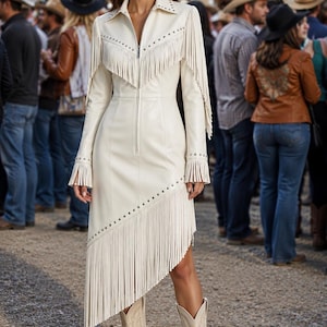 Puede incluir: Un vestido de manga larga en color blanco roto con cierre de cremallera frontal, cuello en punta y adornos de tachuelas plateadas. La prenda incluye detalles de flecos en capas en el pecho, mangas y un dobladillo asimétrico, junto con botas a juego.