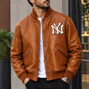 以下が含まれることがあります： 左胸に白と茶色のニューヨーク・ヤンキースのロゴがあしらわれた茶色のレザースタジアムジャンパー。茶色のリブ編みの襟、袖口、裾、フロントのスナップボタン留め、2つのサイドポケットが特徴で、無地の白いTシャツの上に着用されています。