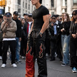 May include: A person wearing a black t-shirt, sunglasses, and black leather pants with flame designs in red and gold. The person is wearing a watch and a silver necklace. The background shows a crowd of people.