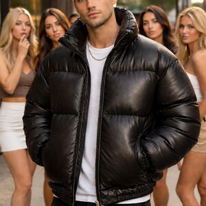 May include: A black puffer jacket with a high collar and a zipper closure. The jacket is worn over a white t-shirt and black pants. A silver chain necklace is visible. Several people are in the background.