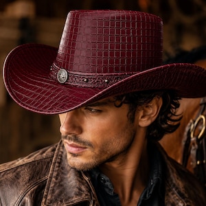 May include: A deep burgundy cowboy hat featuring a textured, grid-like embossed pattern across the crown and brim. The hat is accented with a dark band adorned with small metallic studs and a central circular silver-toned emblem.