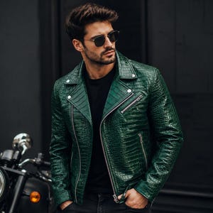 May include: A green leather jacket with a crocodile skin texture. The jacket has a zipper closure and silver hardware. The person is wearing black sunglasses and a black shirt. A motorcycle is partially visible in the background.