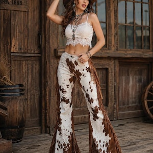 May include: A woman wearing a white lace crop top, a brown hat, and a layered necklace. She is also wearing white pants with a brown cowhide pattern and brown fringe along the sides. The background features a wooden door and a barrel.