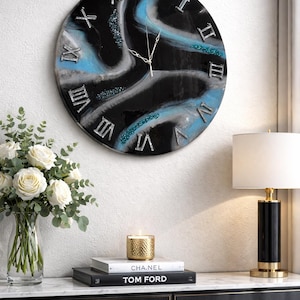 May include: A round wall clock with a black, blue, and silver abstract design. The clock face features Roman numerals and silver hands. The clock is displayed on a white wall, with a table holding books, a candle, and a lamp.