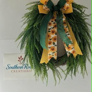 May include: A decorative wreath with green foliage and a large bow. The bow features yellow and green ribbons, with a sunflower pattern on some of the ribbons. The wreath is hanging on a white wall.