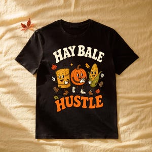 Könnte beinhalten: Ein schwarzes Kurzarm-T-Shirt mit einem Grafikdruck, der den Text "HAY BALE HUSTLE" in Weiß und Orange zeigt. Das Design umfasst Zeichentrickfiguren eines Heuballens, eines Kürbisses und eines Maiskolbens mit Armen und Beinen sowie herbstlichen Blättern.