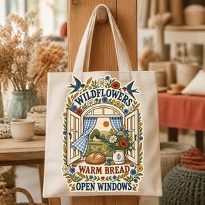 May include: A beige canvas tote bag featuring a colorful graphic of an open window with blue checkered curtains, a loaf of bread, a pitcher of flowers, and two swallows. The text reads Wildflowers, Warm Bread, Open Windows in blue.