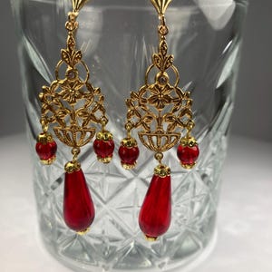 Puede incluir: Pendientes dorados ornamentados con un diseño floral de filigrana. Cada pendiente presenta tres pequeñas cuentas rojas y una gran cuenta roja en forma de lágrima. Los pendientes se exhiben en un vaso transparente.