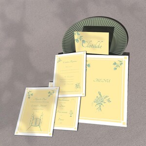 May include: A collection of stationery with a soft yellow background and white borders. The set includes a menu, place cards, and other cards with floral accents and the name "Clotilde". A green plate is in the background.