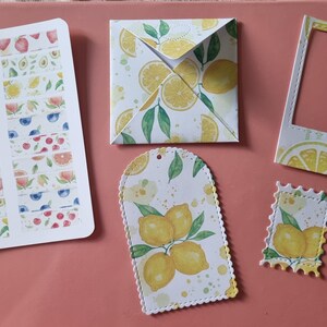 May include: A collection of paper stationery items featuring lemon and fruit motifs. Included are a sticker sheet with various fruit strips, a square folded envelope, a rectangular photo frame, a scalloped tag, and a small postage stamp cutout.