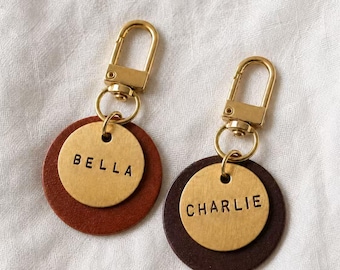 Peut inclure: Deux porte-clés personnalisés pour animaux de compagnie. Chaque porte-clés est doté d'un fermoir et d'un anneau dorés, avec un design superposé d'une base en cuir marron et d'un disque en métal doré. Une étiquette indique "BELLA", l'autre "CHARLIE".