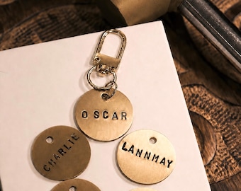Peut inclure: Un ensemble de médailles pour animaux de compagnie personnalisées en laiton, chacune estampillée d'un nom et attachée à un fermoir doré. Les noms sur les étiquettes sont "OSCAR", "CHARLIE", "LANNMAY", "BELLA" et "HATO". Un marteau en laiton est visible en arrière-plan.